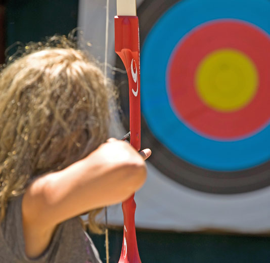 Archery Target inthe background, girl pulling an arrow through the bow